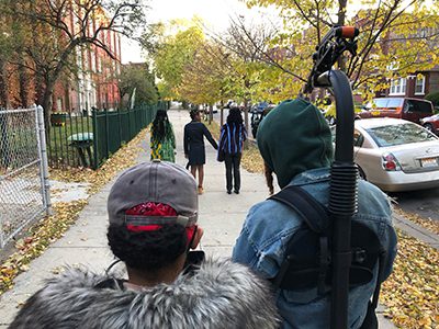 Two people film as 3 young women walk down a sidewalk
